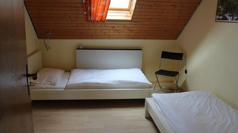 Ein einfaches Zimmer mit zwei Betten und einem Stuhl. Es hat eine Holzdecke und ein Fenster mit einem orangefarbenen Vorhang.