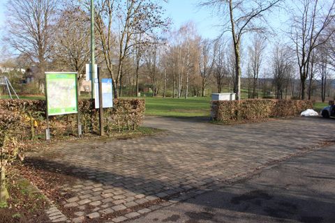 Ein Parkplatz, der an einen Park grenzt, mit Bänken und Schildern. Im Hintergrund sind Bäume und ein klarer Himmel zu sehen.
