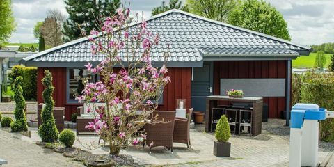 Ein gemütliches Haus mit einem modernen Dach und einem blühenden Baum im Vordergrund. Der Außenbereich ist mit Gartenmöbeln und kleinen Pflanzen gestaltet.