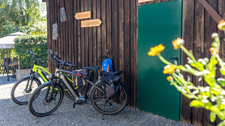 Eine Fahrradgarage aus Holz mit zwei Fahrrädern und einem blauen Rucksack. Im Hintergrund sind Pflanzen und ein Tisch im Freien zu sehen.