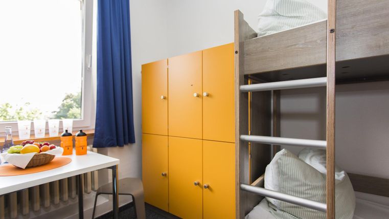 Ein einfaches Zimmer mit einem Hochbett und orangefarbenen Schränken. Auf dem Tisch steht eine Schale mit Obst und Getränke.