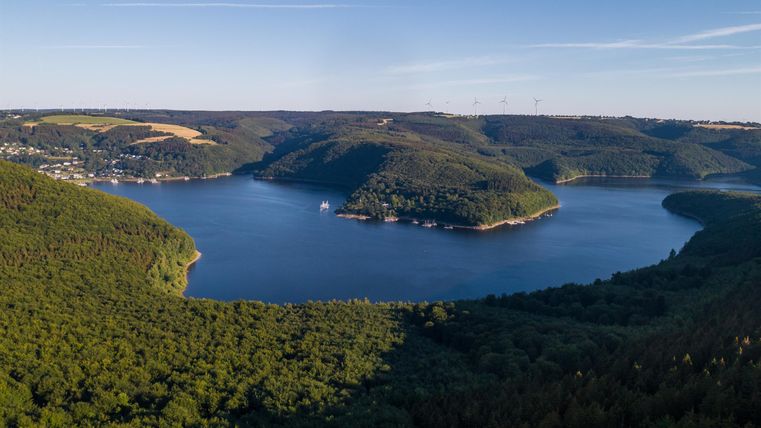 Eine malerische Landschaft mit einem großen See, umgeben von Hügeln und dichtem Wald. Der Himmel ist klar und blau, ideal für Outdoor-Aktivitäten.