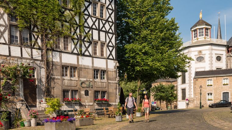 Zwei Personen spazieren auf einer gepflasterten Straße in Kornelimünster, vorbei an einem Fachwerkhaus und einem Café.