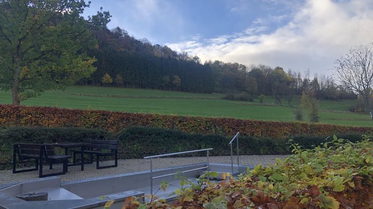 Eine ruhige Landschaft mit einer Aussicht auf grüne Wiesen und Bäume. Im Vordergrund stehen moderne Sitzgelegenheiten auf einem gepflasterten Bereich.
