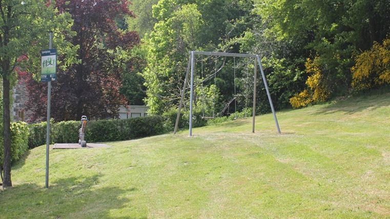 Ein ruhiger Park mit einer Schaukel und üppigem, grünem Gras. Im Hintergrund sind Bäume und ein Busstop zu sehen.