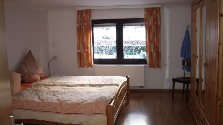 Ein einfaches Schlafzimmer mit einem großen Bett und Holzschrank. Das Zimmer hat ein Fenster mit Vorhängen und einen Stuhl im Hintergrund.