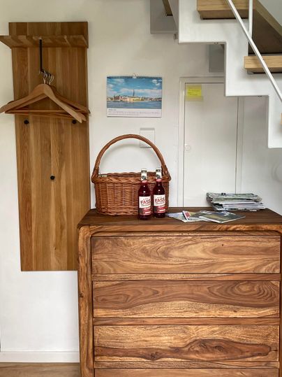 Ein moderner Eingangsbereich mit einer Holzkommode und einem Korb. Im Hintergrund sind eine Garderobe und eine Treppe sichtbar.