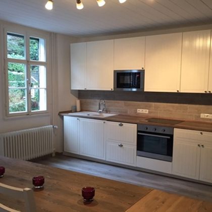 Eine moderne Küche mit weißen Schränken und einem Holzarbeitsbereich. Ein Esstisch mit roten Kerzen steht im Vordergrund und ein Fenster lässt viel natürliches Licht herein.