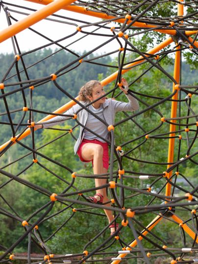 Ein Kind klettert auf einem Klettergerüst aus Seilen. Im Hintergrund sind Bäume und eine grüne Landschaft zu sehen.