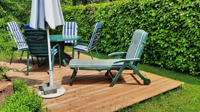 Eine gemütliche Terrasse mit einem Tisch und Stühlen aus Holz. Im Hintergrund ist eine grüne Hecke sichtbar.