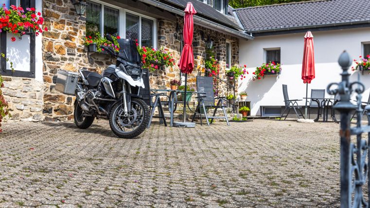 Eine Motorrad steht auf einem gepflasterten Innenhof vor einem schönen Haus. Rote Sonnenschirme und Blumenkästen verleihen der Umgebung eine einladende Atmosphäre.