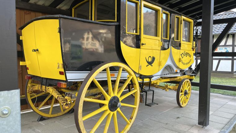 Eine dekorative gelbe Kutsche mit schwarzen Details steht unter einem Dach. Sie hat große, runde Räder und einen nostalgischen Charme.