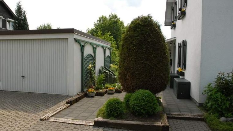 Ein gepflegter Eingang mit einer Hecke und mehreren Pflanzen. Im Hintergrund ist ein schlichter Anbau und ein helles Wohngebäude zu sehen.