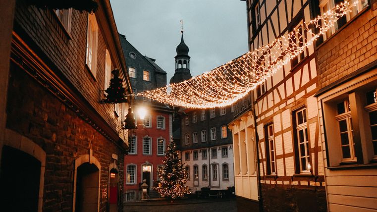 Eine malerische Gasse mit festlichen Lichtern und einem Weihnachtsbaum. Im Hintergrund ist ein Turm zu sehen, der die winterliche Stimmung verstärkt.