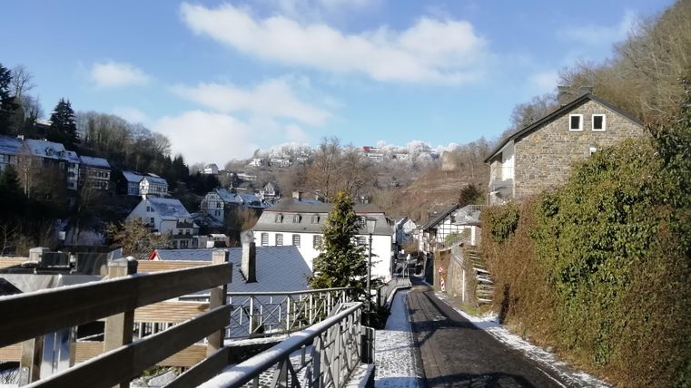 Eine malerische Straße mit Häusern und Bäumen, umgeben von sanften Hügeln. Der Himmel ist klar und sonnig, und der Boden ist mit Schnee bedeckt.