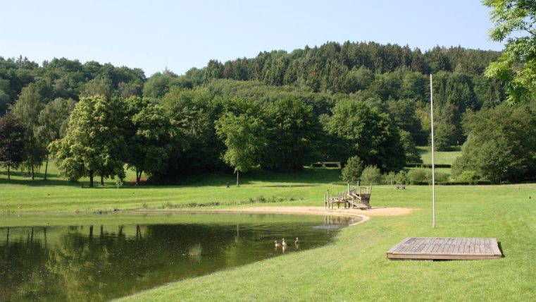 Ein ruhiger Park mit einer Wiese und einem kleinen Teich. Im Hintergrund sind Bäume und Hügel zu sehen.