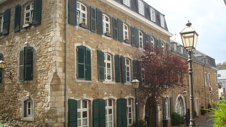 Ein historisches Gebäude mit Steinfassade und grünen Fensterläden. Im Vordergrund geht eine gepflasterte Straße und es sind Laternen zu sehen.