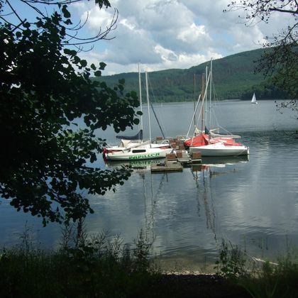 Ein ruhiger See mit mehreren Booten, die sanft im Wasser schwimmen. Umgeben von grünen Bäumen und Hügeln unter einem bewölkten Himmel.