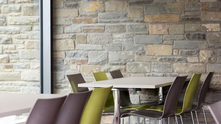 Ein moderner Innenraum mit einem Tisch und farbigen Stühlen. Die Wand besteht aus Naturstein und schafft eine angenehme Atmosphäre.