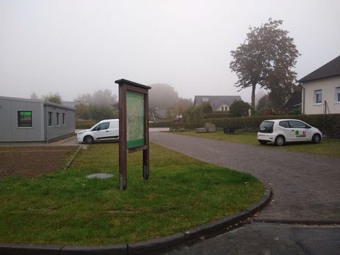 Ein nebliger Ort mit einer Informationstafel und zwei Autos auf dem Parkplatz. Im Hintergrund sind einige Gebäude und Bäume sichtbar.