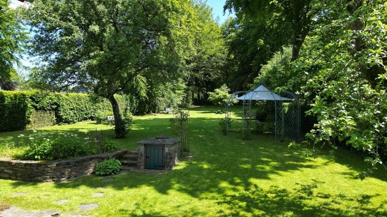 Ein großer, grüner Garten mit viel Rasen und Bäumen. In der Mitte steht ein Pavillon, umgeben von bunten Pflanzen.
