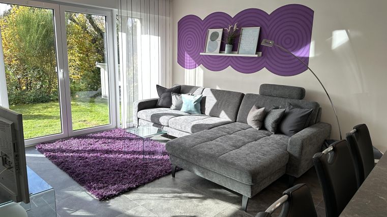 Ein modernes Wohnzimmer mit einem bequemen grauen Sofa und einem lila Teppich. Große Fenster sorgen für viel natürliches Licht und Blick ins Grüne.