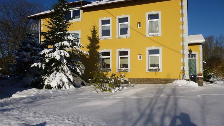 Ein gelbes Haus, umgeben von Schnee. Im Vordergrund stehen schneebedeckte Bäume.