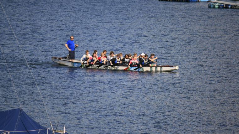 Eine Gruppe von Ruderern in einem Boot auf ruhigem Wasser. Im Hintergrund sind einige Boote und die Uferlandschaft zu sehen.