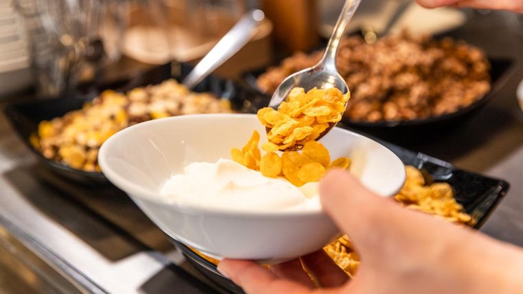 Eine Person füllt Cornflakes in eine Schüssel mit Joghurt. Im Hintergrund sind verschiedene Müsli und Zutaten in Schalen zu sehen.