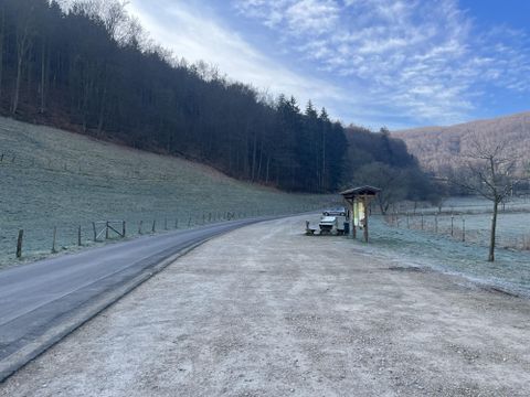 Eine ruhige Landstraße mit Frost auf dem Boden und Bäumen im Hintergrund. Ein kleiner Wartestand befindet sich am Rand der Straße.