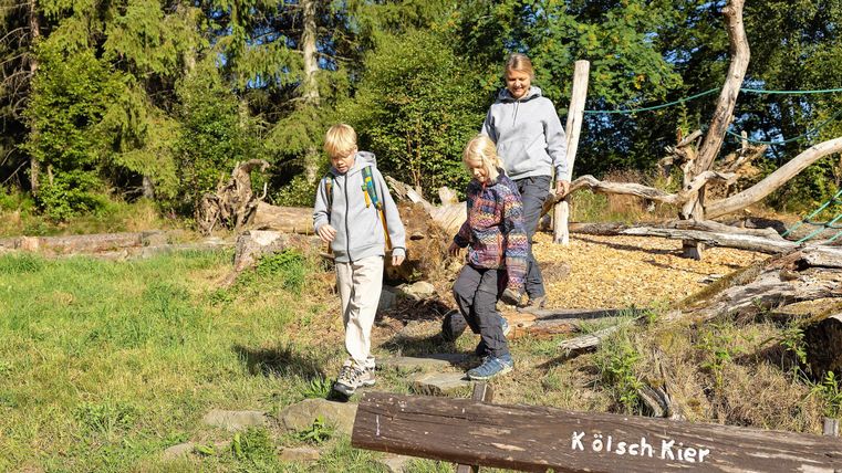 Drei Kinder wandern auf einem Waldweg. Im Vordergrund ist ein Holzschild mit der Aufschrift "Köläch Kier".