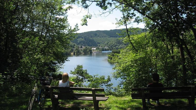 Ein ruhiger See umgeben von Bäumen und Hügeln. Zwei Personen sitzen auf Bänken und genießen die Aussicht.