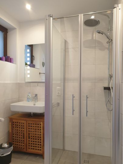 Eine moderne Dusche mit Glastüren und einem Regenbrausekopf. Der Raum hat helle Fliesen und eine stilvolle Waschbeckenanlage.