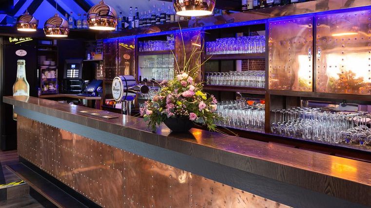 Eine stilvolle Bar mit einer glänzenden Kupferoberfläche und eleganter Beleuchtung. Blumenarrangement und Gläser verleihen der Atmosphäre einen einladenden Touch.
