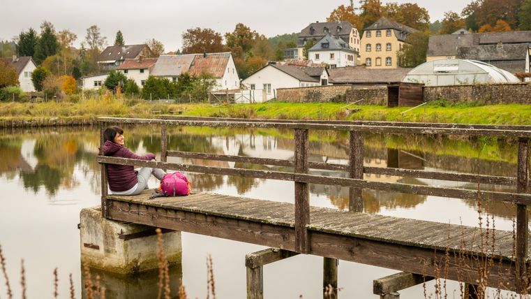 Person sitzt auf einem Steg am Wasser mit Rucksack, im Hintergrund Gebäude und Bäume.