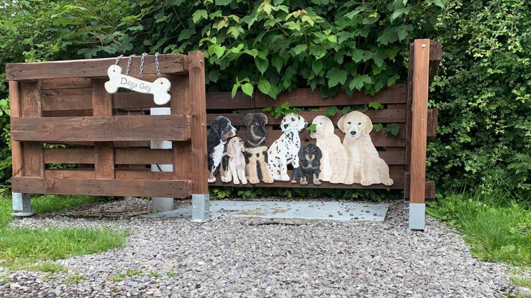 Eine Hundespielstation aus Holz mit verschiedenen Hunde-Silhouetten. Im Hintergrund ist ein grüner Bereich mit Pflanzen zu sehen.