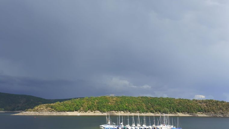 Ein ruhiger See mit mehreren Segelbooten im Vordergrund. Der Himmel ist dunkel und bewölkt, was eine dramatische Stimmung erzeugt.
