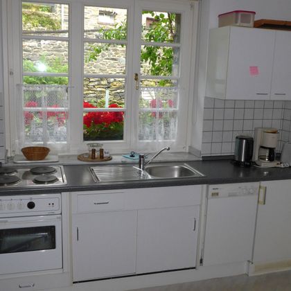 Eine helle Küche mit modernen Geräten und einem Fenster, das auf einen Garten mit Blumenblick zeigt. Die Wände sind in einfachen, hellen Farben gehalten.