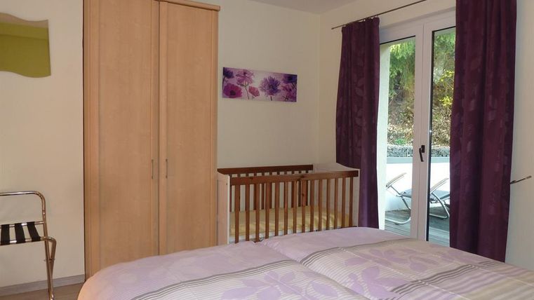 Ein gemütliches Schlafzimmer mit einem Doppelbett und einem Gitterbett. Große Fenster führen zu einem Balkon und die Wände sind in sanften Farben gehalten.