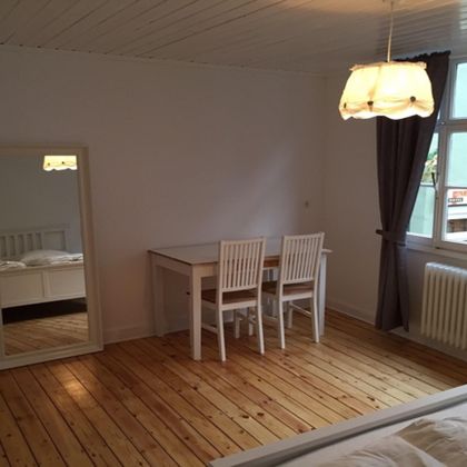 Ein einfaches und helles Zimmer mit einem Tisch und vier Stühlen. Ein großer Spiegel steht an der Wand und die Fenster lassen ausreichend Tageslicht herein.