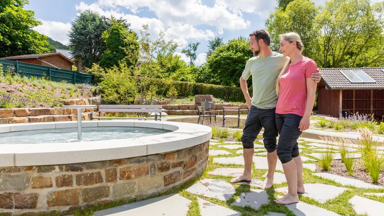 Ein Paar steht entspannt am Rand eines runden Whirlpools in einem schönen Garten. Die Sonne scheint, und im Hintergrund sind Bäume und Gartenmöbel sichtbar.