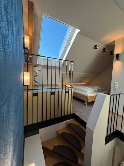 Ein moderner Innenraum mit Holztreppe und hellem Licht. Oben befindet sich ein gemütliches Schlafzimmer mit einem großen Fenster.