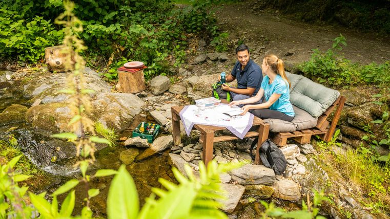 Ein Paar sitzt entspannt an einem Tisch am Flussufer. Sie genießen die Natur und haben eine Karte sowie Getränke dabei.