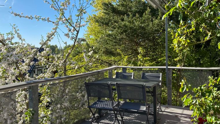 Ein gemütlicher Balkon mit einem Tisch und Stühlen, umgeben von blühenden Bäumen. Die Sonne scheint und der Himmel ist klar.