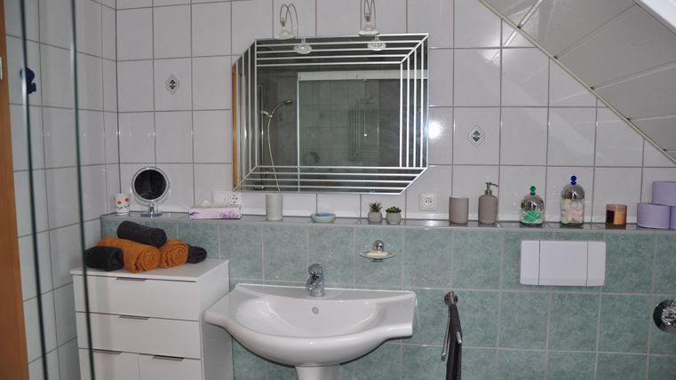 Ein modernes Badezimmer mit grünen Fliesen. Es gibt ein Waschbecken, einen Spiegel und verschiedene Pflegeprodukte auf den Regalen.