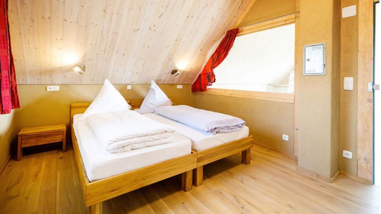 Ein gemütliches Zimmer mit zwei Einzelbetten und Holzfußboden. Die Wände sind hell und die Fenster lassen viel Licht herein.