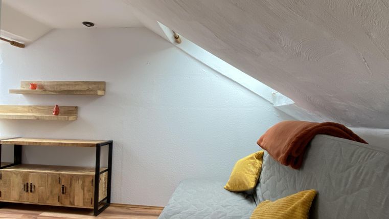 Ein gemütlicher Raum mit einer schrägen Decke, einem Sofa und bunten Kissen. An der Wand befinden sich moderne Regale und ein holzfarbener Schrank.