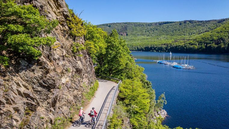 Zwei Radfahrer fahren entlang eines malerischen Ufers. Im Hintergrund sind grüne Hügel und Boote auf dem Wasser zu sehen.