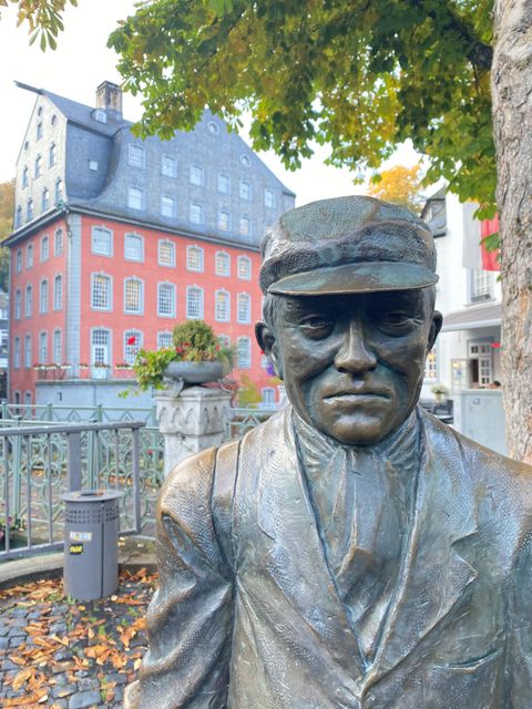 Eine Bronze-Figur eines Mannes in einem Anzug und einer Mütze steht vor einem historischen Gebäude. Im Hintergrund sind weitere alte Gebäude und Bäume zu sehen.
