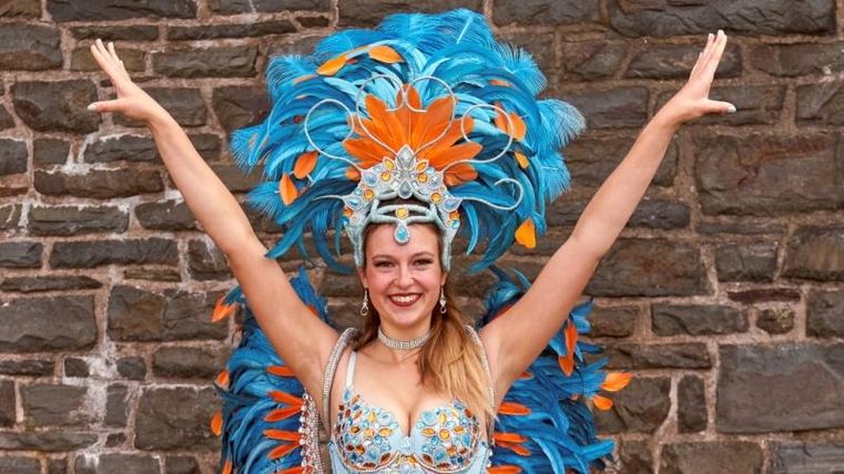 Eine fröhliche Tänzerin mit buntem Federkostüm steht vor einer Steinmauer. Sie hebt die Arme und lächelt, während sie ihre lebhaften Accessoires präsentiert.